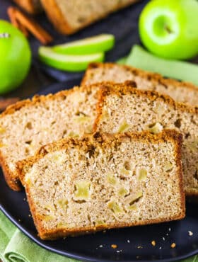 Four slices of Apple Bread on a black plate with whole and cut apples in the background