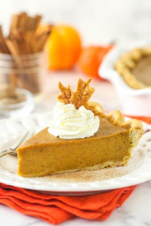 a slice of pumpkin pie on a white plate on an orange napkin with whipped cream and pie crust leaf on top