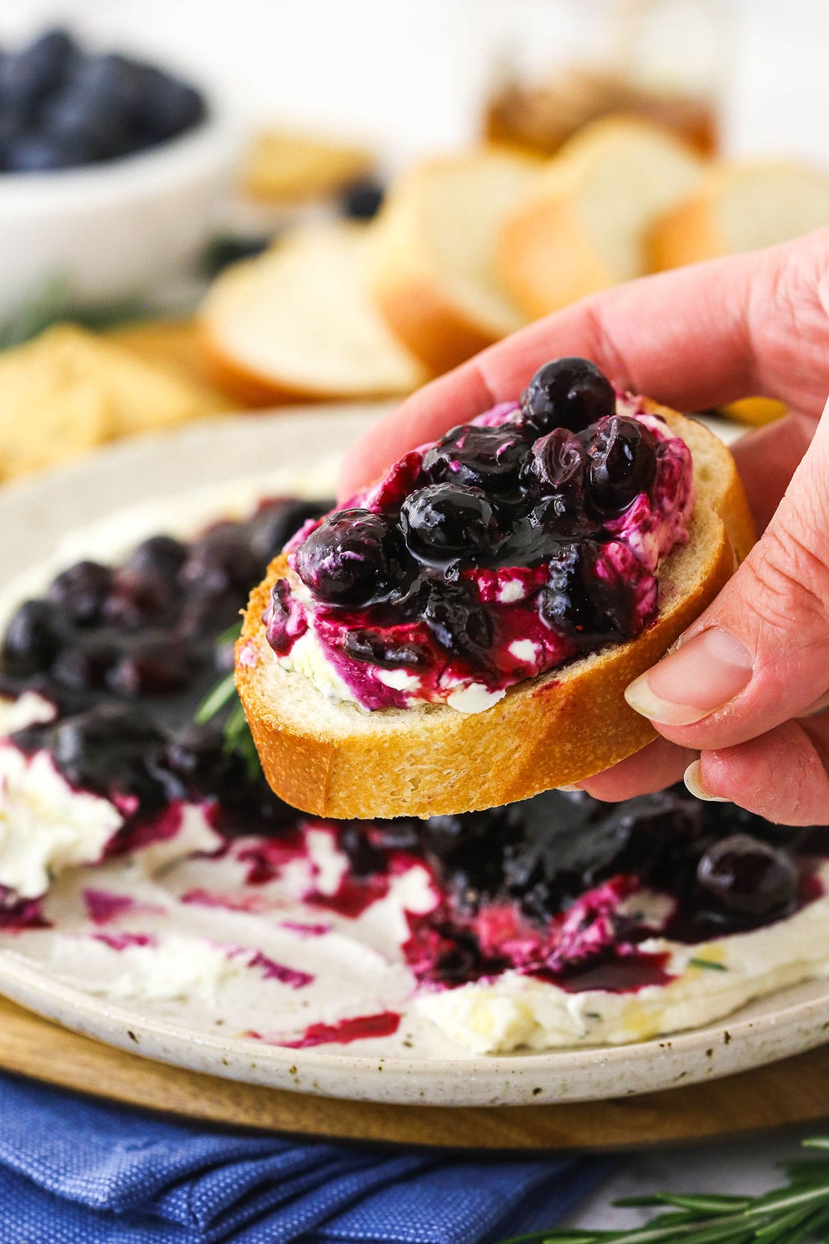 A hand holding a piece of fresh bread topped with blueberry goat cheese dip over a plate full of dip