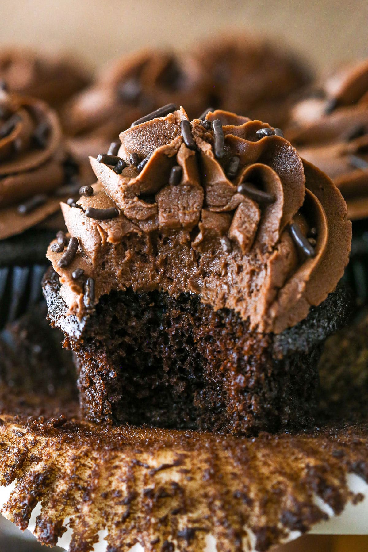 A moist chocolate cupcake with a bite taken out of it alongside other chocolate cupcakes on a cake stand.