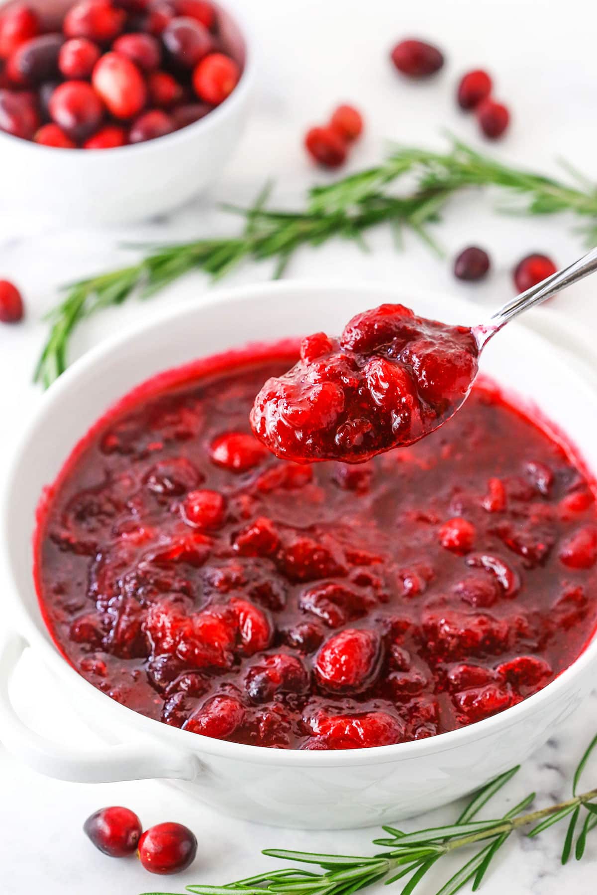 A spoon full of easy homemade Cranberry Sauce over a bowl of Cranberry Sauce in a white bowl on a white table