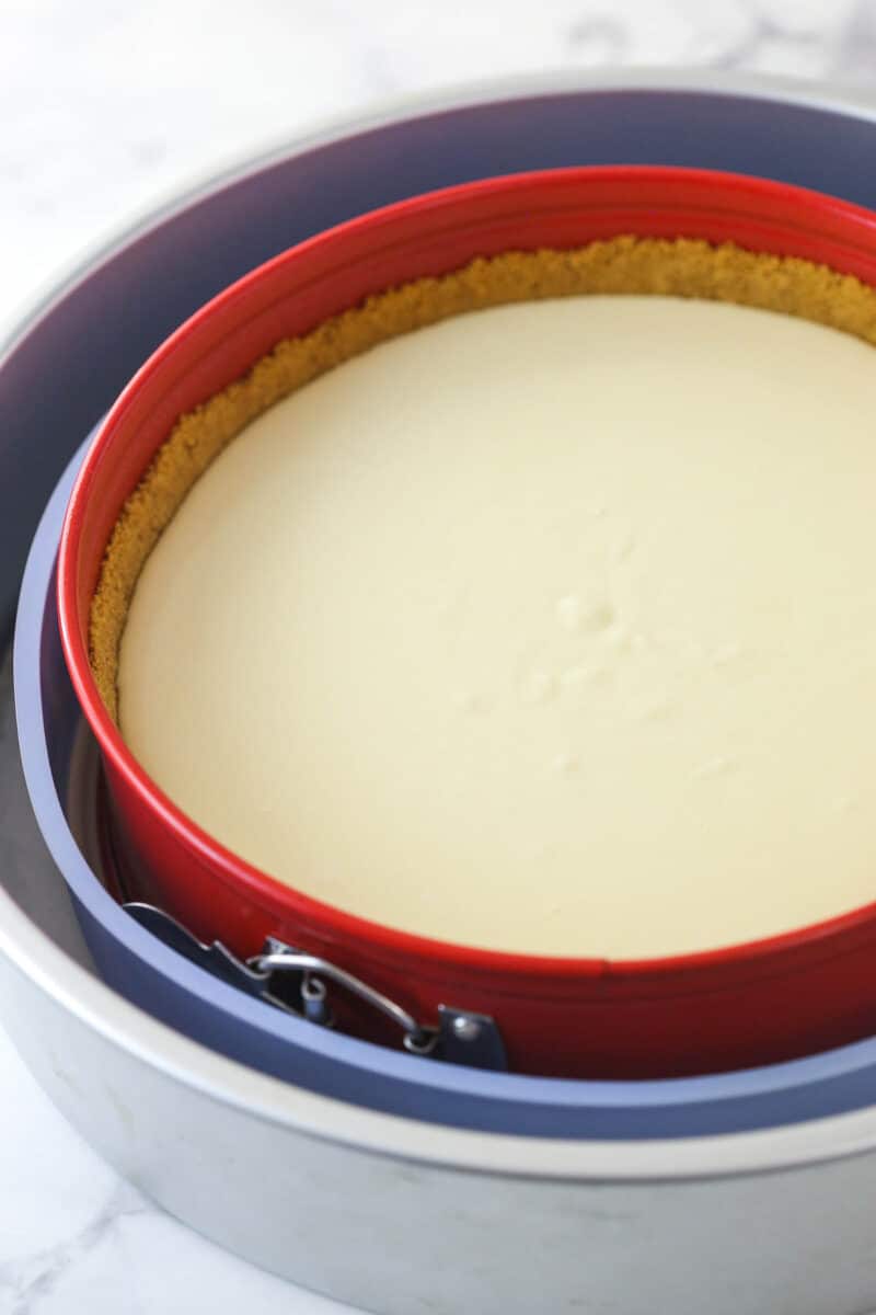 A springform pan in a water bath half full of vanilla cheesecake batter in a graham cracker crust.